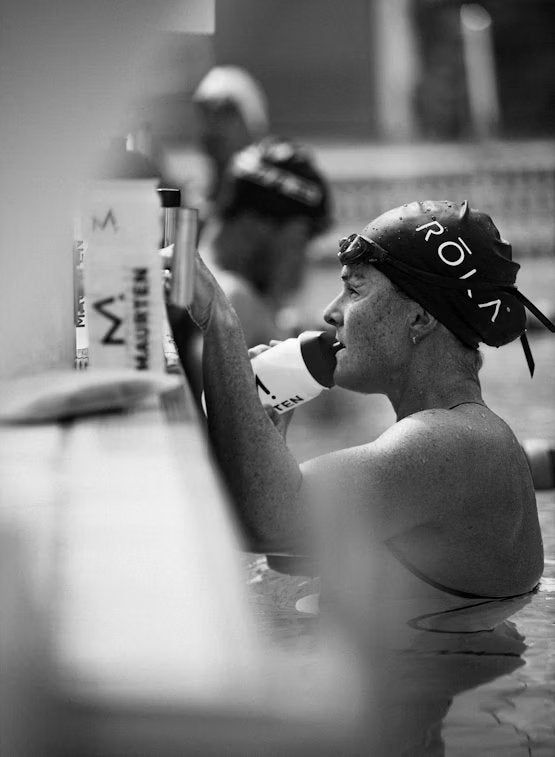 A swimmer wearing a cap drinks from a bottle while resting at the pool edge. Another athlete is blurred in the background. The scene is in black and white. The bottle has a cylindrical shape and smooth surface; specific label text, if present, is not legible. People shown are dressed in standard swim attire. Indoor swimming pool setting with tiled edge visible.