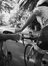 A cyclist grabs a water bottle from someone in a car while riding on a shaded road, surrounded by trees and dappled sunlight. The water bottle is cylindrical, made of semi-transparent plastic with a matte finish. No visible brand or label text can be seen. The road is paved and the background shows leafy trees casting patterned shadows over the scene.