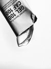 A close-up of a partially opened sports gel packet, with clear gel oozing out against a white background. The packet is rectangular with notched edges, made of glossy flexible material, and features printed black and white text in bold sans-serif font. The label shows GEL 100 CAF 100 and other nutritional information. No people or hands are present.