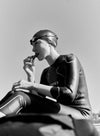 A person in a swim cap and wetsuit sits outdoors, enjoying a snack. The black-and-white image captures the swimmer preparing for or unwinding after an invigorating swim. No visible product packaging or branding; surface finishes appear matte due to lighting. Background shows an outdoor setting with natural elements partially in view. Shot is medium, focusing on the upper body and face of the subject.