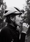 A cyclist wearing a helmet and sportswear eats an energy bar while standing outdoors, surrounded by trees and a cloudy sky. The energy bar is unwrapped, rectangular, and has a textured surface. The wrapper, partially visible in the cyclist’s hand, shows legible printed label text in bold font. Medium shot; no other people are present.