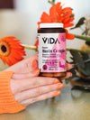 A hand with orange-painted nails holds a cylindrical, opaque white bottle of VIDA Beauty Biotin Complex with a matte finish and a white cap. The label is printed in dark sans serif text and lists biotin, zinc, and essential nutrients. The background shows bright orange flowers filling the frame. This is a close-up shot.