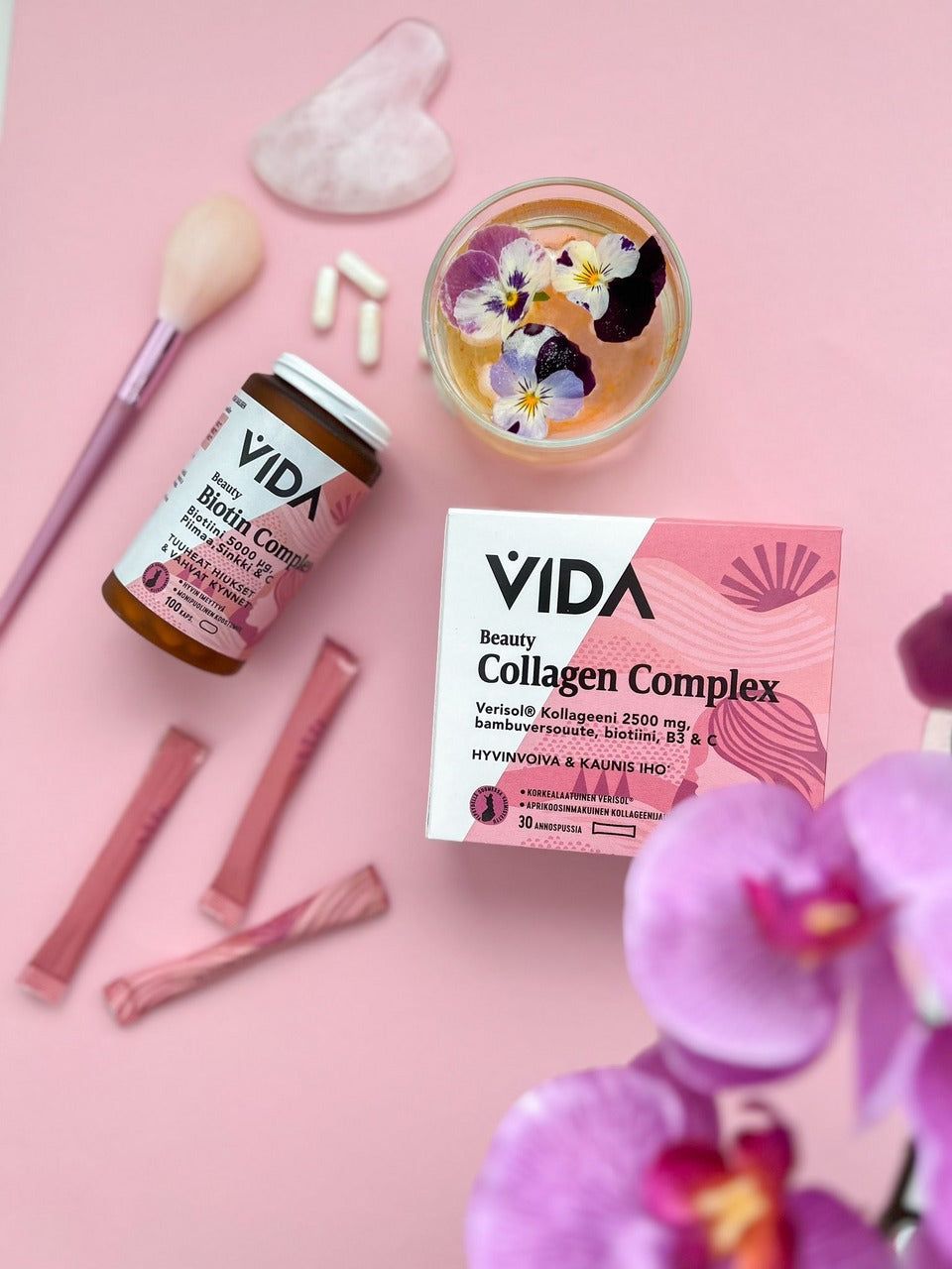 A cylindrical bottle and rectangular box labeled “VIDA Collagen Complex” in sans-serif font, several individually wrapped pink supplement sticks, white capsules, a metallic pink makeup brush with soft bristles, a clear glass vase containing small flowers, and a pink orchid are arranged on a matte pastel pink flat surface. No people or hands are present.