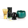 A matcha tea set featuring a bamboo whisk with a natural finish, a black glossy ceramic whisk holder, a matte teal ceramic bowl with round edges, a small canister of premium Moya matcha with green and white packaging and simple sans-serif typography, and a smooth bamboo scoop. All items are neatly arranged on an even white background in an overhead shot.