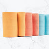Five textured rolls of fabric in orange, pink, and blue stand upright in a row on a white marble surface. The fabrics have patterned surfaces and rounded top edges. No visible packaging or labels. The surface finish is matte with clear texturing visible on each roll. The background is plain white marble. There are no people or hands present. Wide product shot with overhead lighting.