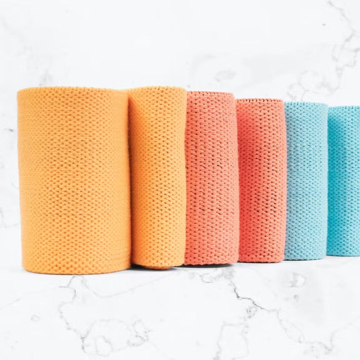 Five textured rolls of fabric in orange, pink, and blue stand upright in a row on a white marble surface. The fabrics have patterned surfaces and rounded top edges. No visible packaging or labels. The surface finish is matte with clear texturing visible on each roll. The background is plain white marble. There are no people or hands present. Wide product shot with overhead lighting.
