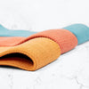A close-up of a rolled, textured yoga mat featuring orange, coral, and teal stripes, placed on a white marble surface. The mat is cylindrical in shape and made from a patterned material with a matte finish. No packaging or typography is visible. There are no people or hands in the image. The background consists solely of smooth white marble.