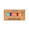 A cardboard box labeled myga Glute Resistance Bands features blue, pink, and orange fabric bands visible through a rectangular plastic window. The box is matte with black sans-serif text and simple outline illustrations of people using resistance bands. The packaging is rectangular, primarily in neutral tones. No people or hands are present. Plain white background, product photographed straight-on at eye level. Outline drawings of exercise poses and resistance band exercises are printed on the box.