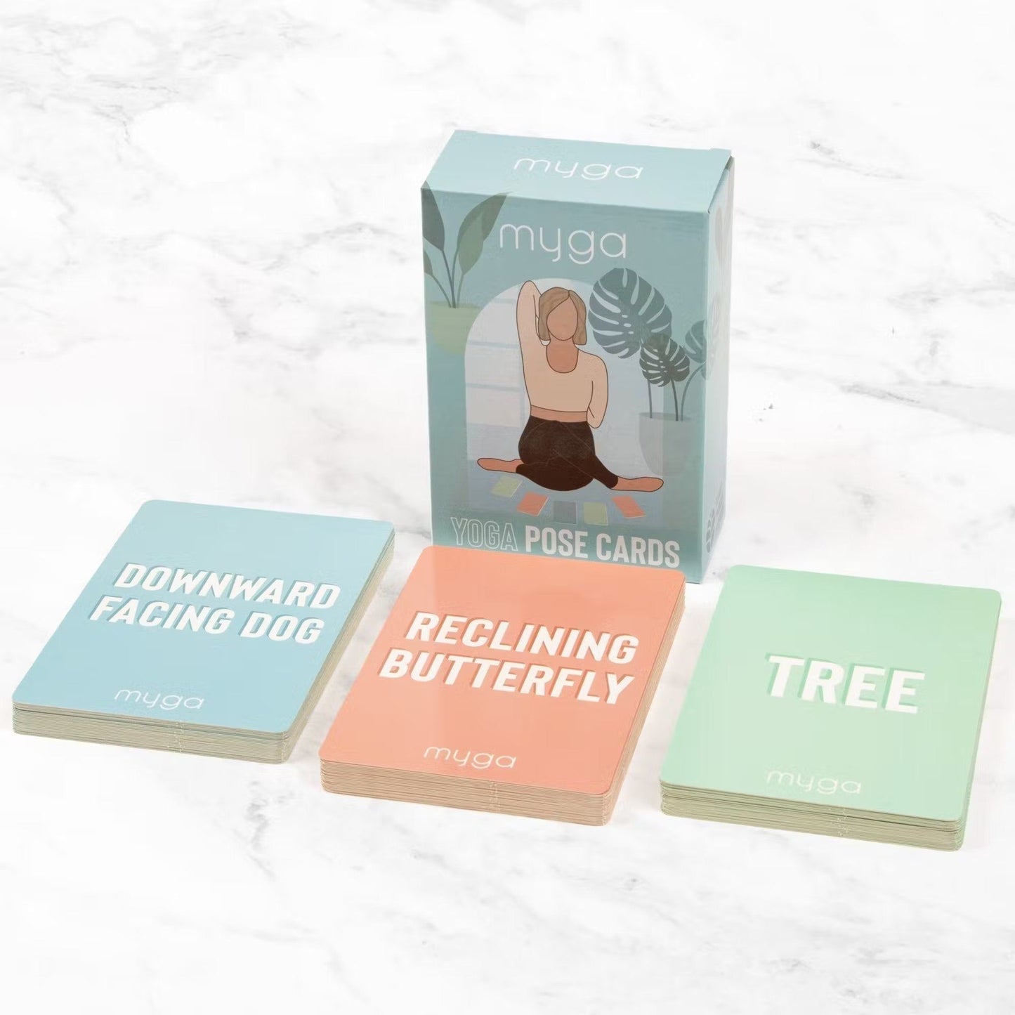 Myga Jooga Asentokortit; laatikko Myga Yoga Pose Cards -korteille kolmen kortin kera edessä – Downward Facing Dog, Reclining Butterfly ja Tree – valkoisella marmoripinnalla; laatikko suorakulmainen pahvia mattapinnalla; kussakin kortissa kuvitettu joogaliike ja liikkeen nimi selkeällä groteskitekstillä; ei ihmisiä tai käsiä, kuva ylhäältä kuvioidulla kivipinnalla.