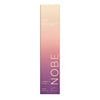 A tall, rectangular box for NOBE Oat Wonder Luminous Lip Oil features a vertical gradient, shifting from light beige at the top to a deep purple at the bottom, with gold-toned text throughout. The box is made of matte-finish cardboard and has sharp edges. “NOBE” is printed vertically in sans-serif font. Oat imagery and product details are included on the front. No people or hands are visible; plain background; studio shot.