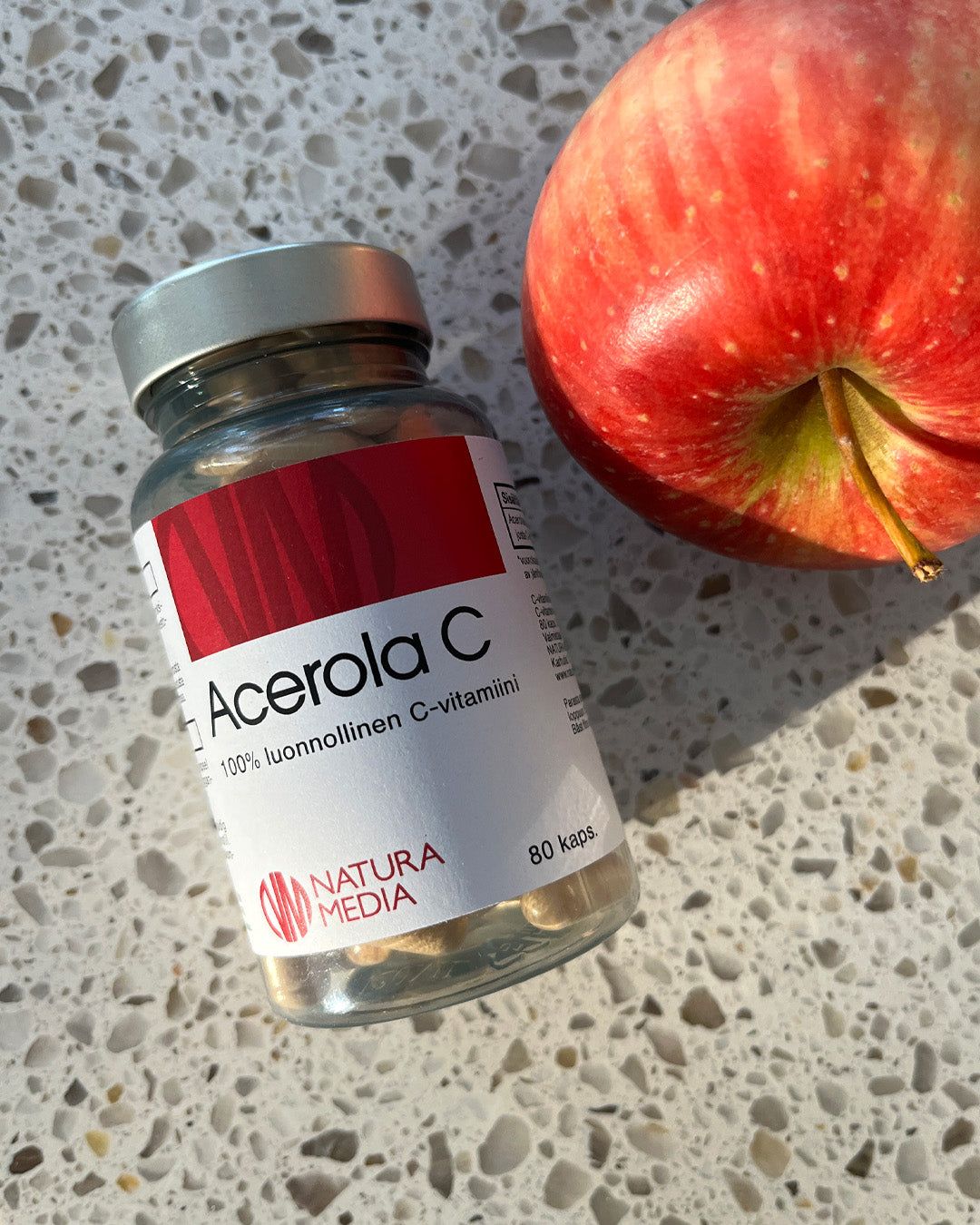 A white plastic bottle with a matte finish labeled “Acerola C” and a red apple rest on a light-colored, speckled countertop in direct sunlight. The bottle has a white cap and cylindrical shape. Label text states “80 capsules, natural vitamin C by Natura Media” with acerola extract benefits. No people or hands are present. A bottle labeled Acerola C and a red apple rest on a speckled countertop in sunlight. The bottle contains 80 capsules of natural vitamin C by Natura Media, highlighting the benefits of