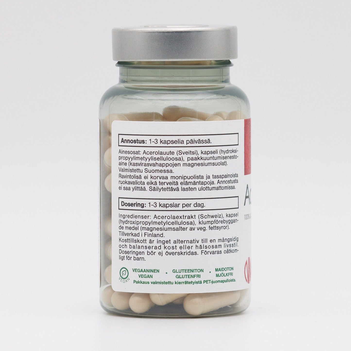 A glass bottle of dietary supplement capsules with a cylindrical shape and clear, glossy surface. The bottle has a screw-top cap. Its printed label in Finnish and Swedish shows dosage instructions, ingredients, and features small vegan, gluten-free, and lactose-free icons. Modern sans-serif typography is used. The background is plain white. No people or hands are present in the image.