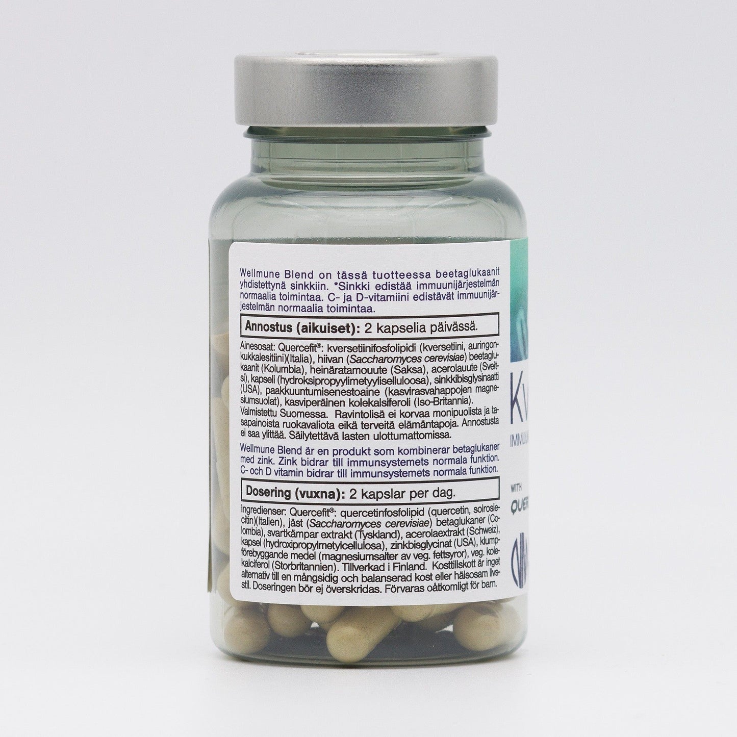 A clear cylindrical bottle with a reflective silver screw-on lid contains multiple beige capsules. The bottle’s white label features black sans-serif text presenting supplement ingredients and dosage instructions in Finnish, Swedish, and English. Product is photographed upright on a plain white background. A clear bottle with a silver lid containing capsules. The label on the bottle lists supplement ingredients and dosage instructions in Finnish, Swedish, and English. The beige capsules inside are