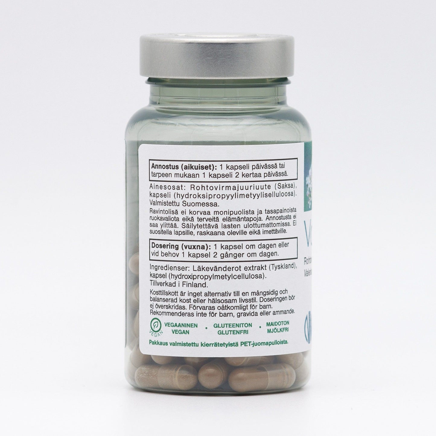 A clear plastic bottle containing herbal supplement capsules, with a cylindrical shape and flat white screw cap. The surface is glossy. The white label features black and green sans-serif text listing ingredients and dosage instructions in both Finnish and Swedish, plus vegan, gluten-free, and dairy-free icons. The product sits on a neutral background. A clear bottle of herbal supplement capsules with a white label listing ingredients and dosage instructions in Finnish and Swedish. The label also displays