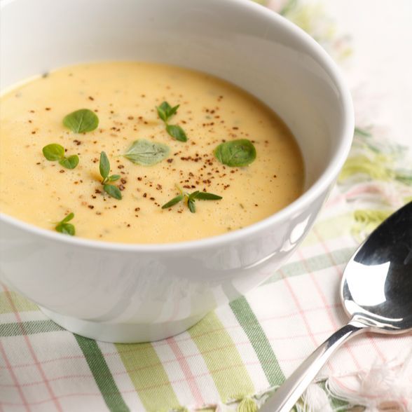 Nutrilett VLCD Kanakeitto; valkoinen kulhollinen kurkumapohjaista, kermaisen keltaista keittoa vihrein yrtein ja mustapippurilla koristeltuna, ruutukuvioisen liinan päällä metallilusikan vieressä; keraaminen ja mattapintainen pyöreä kulho; ruutukangas punaisin ja sinisin sävyin; ei pakkausta, tekstiä tai ihmisiä; kuvattu ylhäältä.
