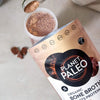 Planet Paleo Chocolate -pussi yläviistosta, edessä vaahtoava suklaajuoma lasissa ja jauhetta valumassa mittalusikasta