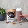 Planet Paleo Chocolate -pussi pöydällä kasvin vieressä, käsi pitelee shakeria suklaanruskealla juomalla