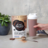 Planet Paleo Chocolate -pussi pöydällä kasvin vieressä, käsi pitelee shakeria suklaanruskealla juomalla