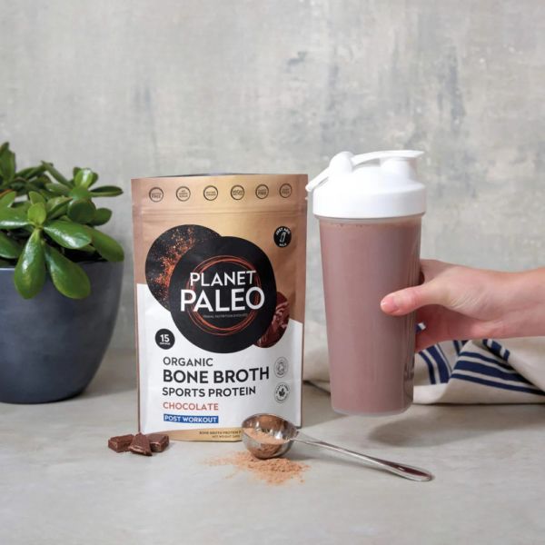 Planet Paleo Chocolate -pussi pöydällä kasvin vieressä, käsi pitelee shakeria suklaanruskealla juomalla