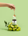 A hand holds a gold spoon above a cylindrical jar labeled Daily Elektrolyyttii. The jar is matte white with a minimal label in black sans-serif font. Several whole and sliced limes are arranged around the jar on a smooth, light green background. The spoon contains fine, bright green powder from the jar. Shot is close-up and well-lit.