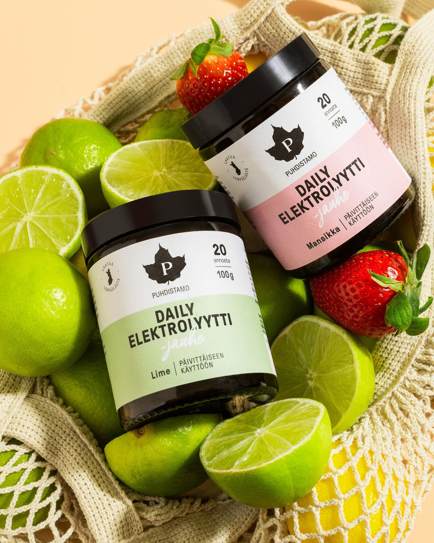 Two jars of Puhdistamo Daily Elektrolyytti powders (Lime and Strawberry) rest on a mesh bag, surrounded by fresh limes and strawberries, displayed on a light yellow surface. The jars are cylindrical with matte white plastic bodies and flat white lids. Black sans-serif branding is printed vertically; each jar has colored labels reflecting the flavor. No people or hands visible.