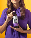 A woman in a purple shirt holds a transparent, cylindrical glass bottle with a matte label displaying “Kombucha” and the flavors “Goji” and “Passionfruit” in sans-serif type. The liquid inside the bottle is amber-colored. The background is solid bright yellow. Medium close-up shot, focus on the bottle and woman’s upper body.