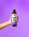 A hand holds a cylindrical, transparent bottle containing purple kombucha. The label displays “KOMBUCHA Cool & Passion” in large, sans-serif white lettering. The bottle has a silver cap and smooth surface with a glossy finish. The background is solid purple with no additional objects or textures visible. Shot is front-facing and closely cropped.