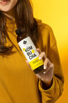A woman in a yellow sweatshirt holds a clear glass bottle containing kombucha. The bottle has a cylindrical shape and features a yellow and white label with bold, modern typography; brief legible text includes the product name. The label finish is matte. Background is solid bright yellow with evenly lit lighting.