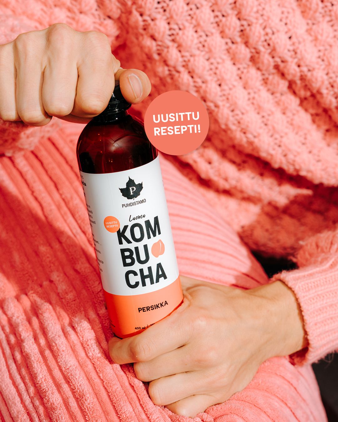 Person in a pink sweater holds a clear glass bottle of Puhdistamo kombucha, peach flavor. The bottle has a matte label showing product name, brand logo, and the text “UUSITTU RESEPTI!!” in bold font. Background is bright solid pink. Only the person’s hand and part of their torso are visible in the shot.