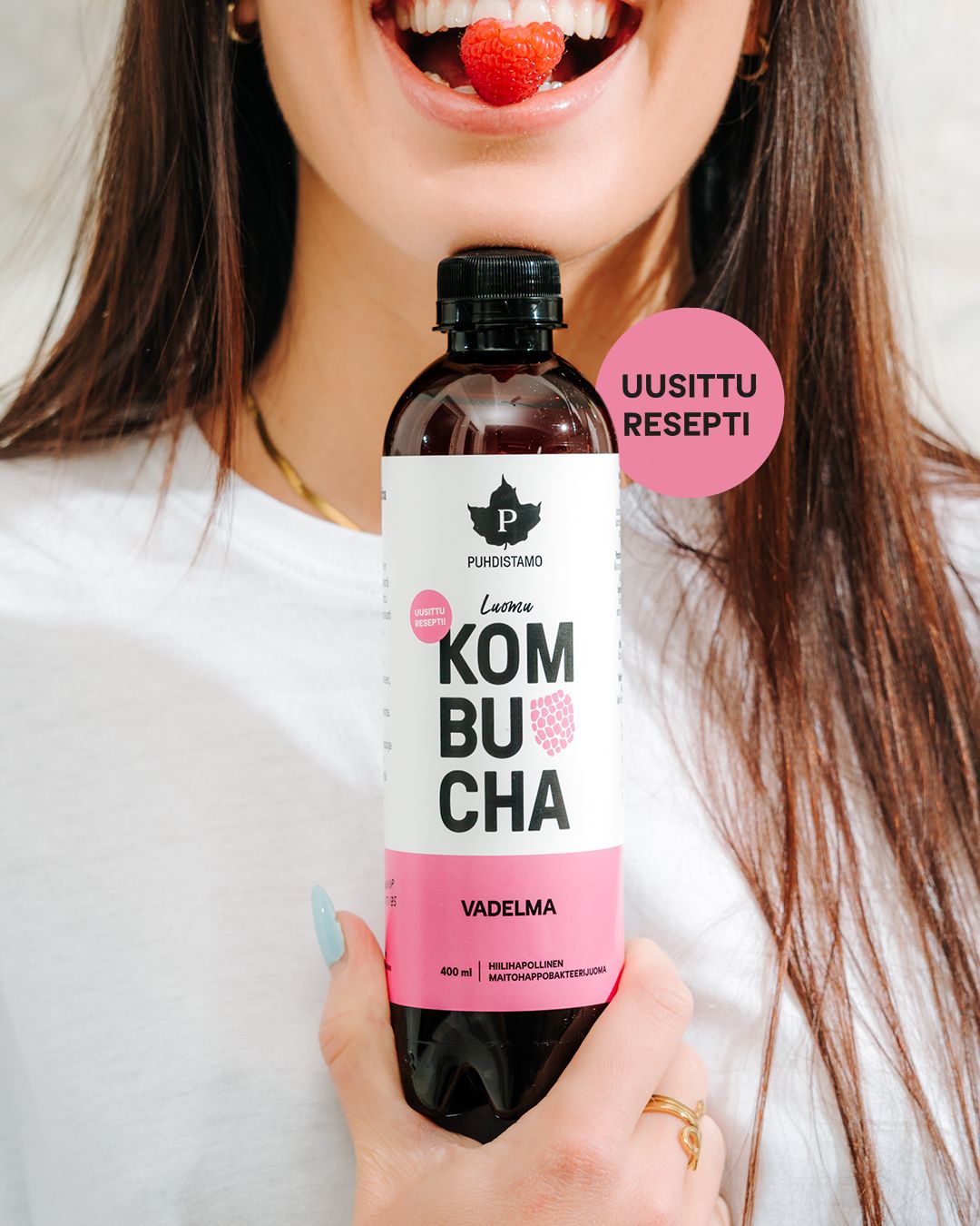 A woman holds a clear glass bottle filled with pink liquid, labeled as raspberry kombucha. The label features bold uppercase sans-serif text reading “KOMBUCHA VADELMA” and a pink circle with “UUSITTU RESEPTI.” The bottle has a matte paper label. She smiles with a raspberry held between her teeth. The background is neutral and uncluttered.