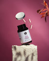 A supplement bottle with a removed lid and three pink capsules floating above a textured stone block. The bottle is cylindrical, made of clear or lightly colored plastic, and has a screw-top lid placed next to it. No visible people or hands. The background is solid pink with several blurred orange leaves on the right side. No legible label text is visible on the bottle.