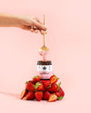 A hand sprinkles powder with a spoon into a clear cylindrical jar labeled “Daily Elektrolyytti” in bold sans-serif type. The jar sits on a matte surface, surrounded by a pile of whole strawberries and loose white electrolyte powder. Pale pink background. Product and ingredients are arranged in the foreground; no other objects are visible.