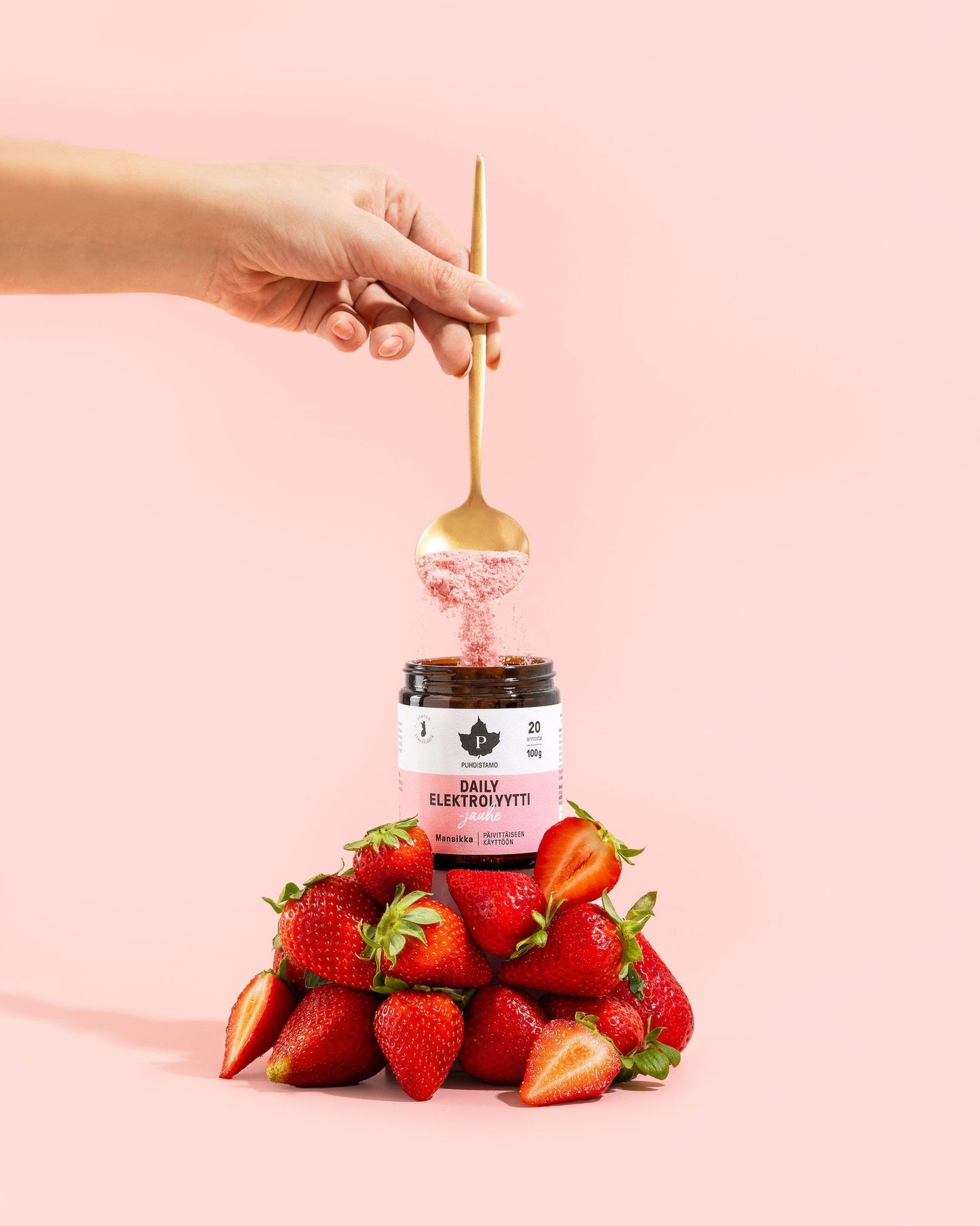 A hand sprinkles powder with a spoon into a clear cylindrical jar labeled “Daily Elektrolyytti” in bold sans-serif type. The jar sits on a matte surface, surrounded by a pile of whole strawberries and loose white electrolyte powder. Pale pink background. Product and ingredients are arranged in the foreground; no other objects are visible.