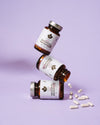 Three brown supplement bottles with white matte labels are stacked at angles, forming a sculptural arrangement. The bottles have cylindrical shapes with smooth surfaces and dark caps. Several white capsules are spread on the flat surface close to the bottles. Label typography is modern and sans-serif, partly visible but not fully readable in the photo. Background is a solid light purple color without texture or props. No people or hands present; shot is still life, frontal perspective showing all products