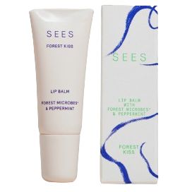 A tube of SEES Forest Kiss lip balm stands next to its box. Both items have a matte finish, with packaging decorated by blue and green abstract lines and simple sans-serif text. The label reads that the balm is infused with forest microbes and peppermint. The shot uses a plain background, no people or hands are present.