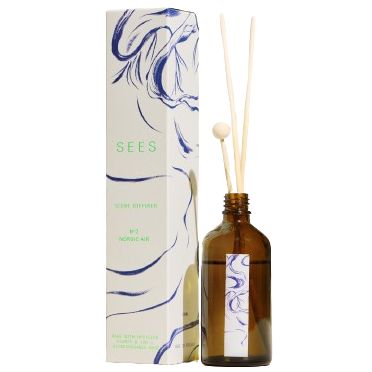 Brown glass reed diffuser bottle with wooden sticks beside a white rectangular box. The box has a matte surface, blue abstract art on the front, and green uppercase SEES lettering. No visible branding on the bottle or sticks. Set against a plain background with no people present. Shot is taken from the front at eye level.
