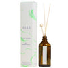 A brown glass bottle with reed diffuser sticks stands beside a white and green SEES box featuring the Nordic Bloom label. The diffuser bottle is cylindrical with a matte finish and several natural-colored wooden sticks inserted. The SEES box has block sans-serif lettering, color blocking in white and forest green, and sharp edges. Both items are positioned on a plain light surface; background is off-white. No people or hands are present in the image.