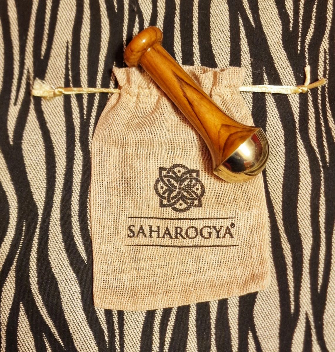 A wooden and brass massage tool rests on a small drawstring pouch labeled SAHAROGYA with a decorative logo, set against a zebra-patterned, holistic fabric background. The massage tool has light wood with glossy brass ends and is cylindrical in shape. The pouch is off-white canvas with bold sans-serif text. No people or hands are present; this is an overhead product shot.