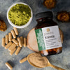 Saharogya Karela 500 mg; pullo valkoisella muovirungolla ja vihreällä korkilla, vihreä-mustaa painettua etikettitekstiä; asetettu mattapintaiselle tekstuuripinnalle pienen vihreää hienojauhetta sisältävän kulhon, irtokapseleiden, viipaloidun tuoreen karelan ja metallilusikallisen karelajauheen kera; ei ihmisiä tai käsiä.