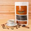 A cup of tea on a white saucer is placed next to a rectangular paper package labeled “Saharogya Luxury Tea Peach Detox” in printed bold serif and script fonts. Loose tea leaves and dried petals are spread on the light wood surface. The background is also light wood. No people or hands are present. Medium product shot, straight-on angle.