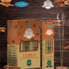 Two boxes of Ullas Orgánico canela (cinnamon) incense sticks are displayed vertically, with two incense sticks burning beside them. Rectangular cardboard boxes feature a matte finish and a brown color palette; packaging includes printed images of cinnamon sticks and white sans-serif label text with green elements. Cinnamon sticks and brand logos enhance the natural atmosphere in the background. No people or hands visible. The setting is a flat tabletop surface photographed from above at a slight angle.