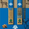 Two boxes of organic incense sticks (White Musk and Almizcle Blanco) are displayed upright on a blue and tan background, featuring deer illustrations and hazelnuts. Rectangular cardboard boxes have matte surfaces with distinct label fonts; one uses thin sans-serif, the other bold serif. Both feature prominent green and white certification seals for organic products. No people or hands visible in the shot.