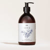 SEES x Moomin Hand Wash; ruskea muovinen pumppupullo mattapinnalla ja pyöristetyllä muodolla; etiketissä lukee SEES Hand Wash, näyttää yksinkertaisia viivapiirroksia kasveista ja muumihahmokuvituksen; typografia selkeää groteskia; lisätekstissä tuoksu: neulametsä setripuulla ja bergamotilla; tausta yksinkertainen ilman näkyviä käsiä tai rekvisiittaa.