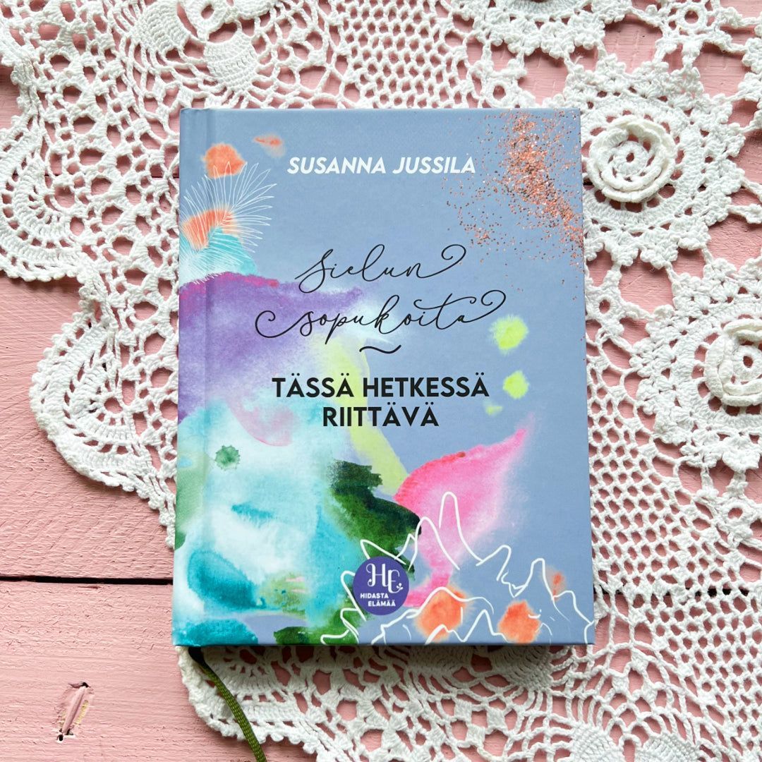 Hidasta Elämää Sielun Sopukoita Lahjaboksi; värikäs kirja tekstillä "Sielun Sopu Kohta Tässä Hetkessä Riittävä" kirjoittajalta Susanna Jussila, aseteltuna vaaleanpunaiselle puualustalle, hento virkattu liina osittain näkyvissä alla; kirjan kansi mattapintainen ja monivärinen lihavoidulla typografialla etusivulla; kuva otettu ylhäältä, ei ihmisiä tai käsiä.