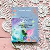 A colorful book titled Sielun Sopu Kohta Tässä Hetkessä Riittävä by Susanna Jussila rests on a pink wooden surface, with a delicate crocheted doily partially visible underneath. The book has a matte cover finish and displays bold, multicolor typography on the front. The shot is overhead with no people or hands shown.