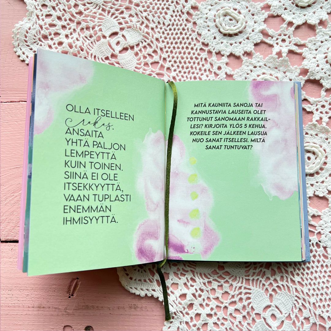 An open book with pale green, matte-finished pages and purple watercolor accents lies flat on a white lace doily atop a pink wooden table. Both left and right pages have printed blocks of text in neat, sans-serif typography; the left side is mostly Finnish prose, the right includes lines for writing. No people or hands are present. An open book with pale green pages and purple watercolor accents rests on a white lace doily and a pink wooden table. The left page features text in Finnish about
