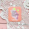 A pink coaster with Finnish text rests on a lacy white tablecloth and a pink wooden surface. The coaster is round, appears to be made of a rigid material with a matte finish, and displays soft pastel-colored artwork. The visible Finnish text uses clear sans serif typography. No people, hands, or packaging are present in the shot.