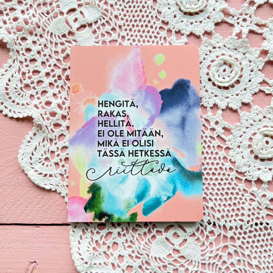 Hidasta Elämää Sielun Sopukoita Lahjaboksi; vaaleanpunainen kortti suomenkielisellä tekstillä, eloisin vesiväritehostein ja tyylikkäällä käsialakirjoituksella, aseteltuna vaaleanpunaiselle puupinnalle valkoisin virkatuin pitsiliinoin; kortti suorakulmainen mattapintaista pahvia, monivärisiä vesiväriaiheita; typografia yhdistää painettua suomenkielistä tekstiä ja koristeellista käsialaa; ei pakkausta tai ihmisiä näkyvissä.