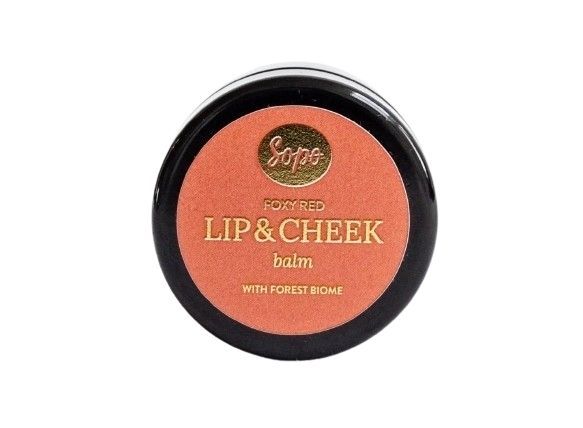 Sopo Sävyttävä Metsäbiomi Huulivoide & Poskipuna; pyöreä rasia tekstillä "Sopo Foxy Red Lip & Cheek Balm" ja maininnalla Forest Biome oranssilla taustalla, kuvattu ylhäältä; rasia pyöreä mattapinnalla, tuotenimi selkeällä groteskitypografialla; ei ihmisiä eikä käsiä näkyvissä; tausta yhtenäisen oranssi, kuva otettu ylhäältä.