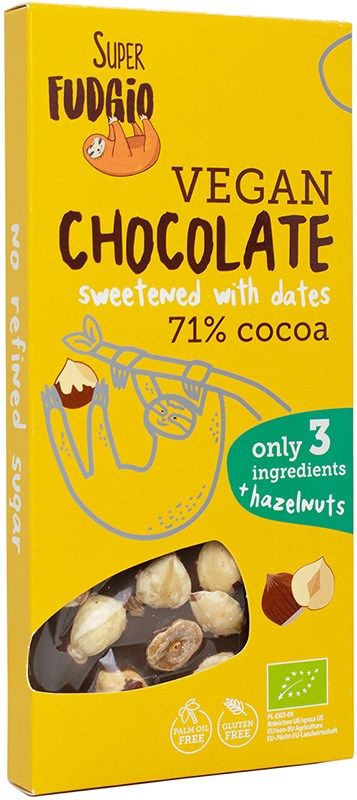 A yellow box of Super Fudgio vegan chocolate with hazelnuts, sweetened with dates, featuring 71% cocoa and made with only three ingredients. Cardboard packaging is rectangular, matte finish. Sans-serif font used for product name and features. Graphics include cocoa pods and hazelnuts illustration. Text highlights no refined sugar, palm oil free, gluten free. Plain background; no people present.